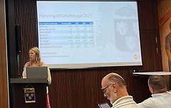 Bild från kommunfullmäktige där en kvinna står i i talarstolen och pratar. På skärmen i bakgrunden ses information och siffror om budget.