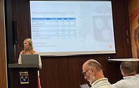 Bild från kommunfullmäktige där en kvinna står i i talarstolen och pratar. På skärmen i bakgrunden ses information och siffror om budget.