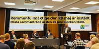 Bild från fullmäktige där en man står i talarstolen och pratar. Ledamöter lyssnar. Med svart text på gul bakgrund står information skriven att kommunfullmäktige 28 maj är inställt.