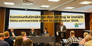 Bild från fullmäktige där en man står i talarstolen och pratar. Ledamöter lyssnar. Med svart text på gul bakgrund står information skriven att kommunfullmäktige 28 maj är inställt.