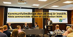 Bild från fullmäktige där en man står i talarstolen och pratar. Ledamöter lyssnar. Med svart text på gul bakgrund står information skriven att kommunfullmäktige 28 maj är inställt.
